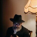 Elderly man in hat using smartphone, sitting by vintage lamp indoors.