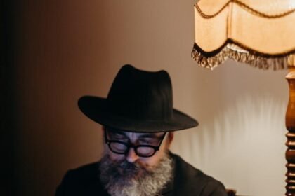 Elderly man in hat using smartphone, sitting by vintage lamp indoors.