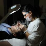 A beautician provides professional eyebrow care to a client in a salon, under focused lighting.